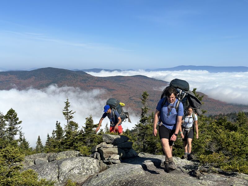 The image shows three hikers on a rocky mountain trail. They are equipped with large backpacks, suggesting a multi-day trip. The background features a scenic view of mountains partially obscured by clouds or fog, creating a sense of depth and altitude. The sky is clear and blue, indicating good weather conditions for hiking.